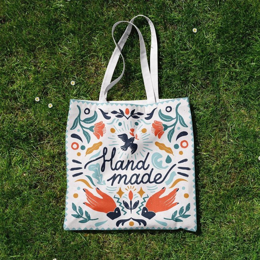 Tote bag mockup featuring a white fabric bag with colorful hand-drawn floral and bird illustrations and 'hand made' text, photographed on green grass.