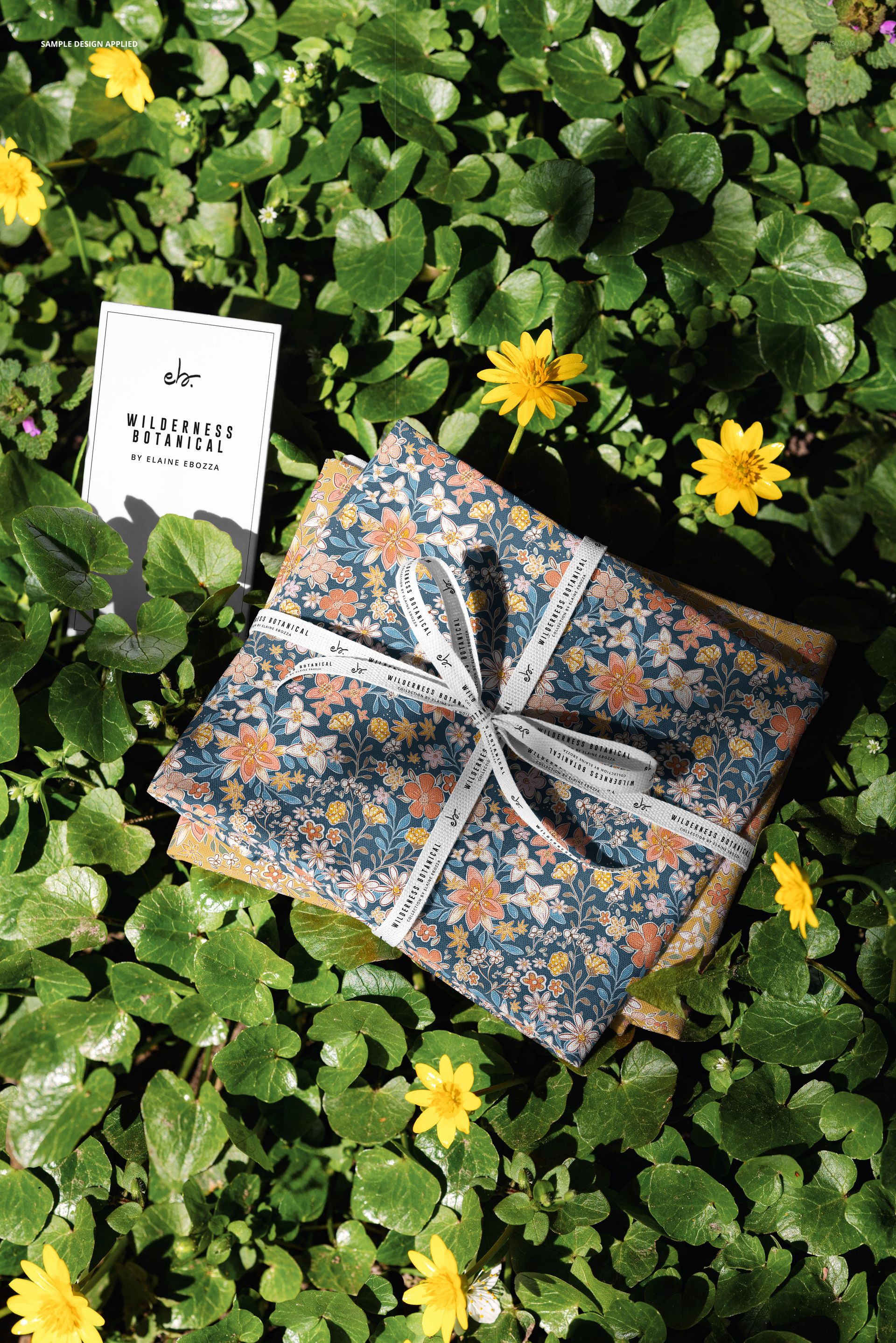 Floral patterned fabrics tied with ribbon placed on leafy ground with scattered yellow flowers, PSD mockup scene.