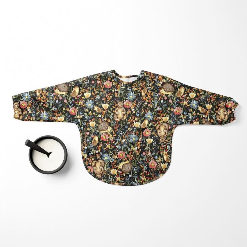 Baby sleeved bib mockup featuring a dark floral and woodland animal pattern, laid flat on a white surface next to a cup of milk.