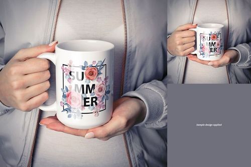 White ceramic mug with floral design and bold text, held in both hands by a person wearing a light gray jacket, front view.