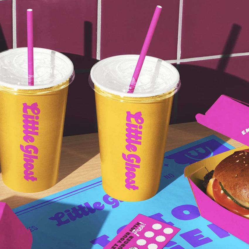 Burger branding packaging mockup featuring two yellow cups with pink straws, a burger box, and a burger on a colorful surface.