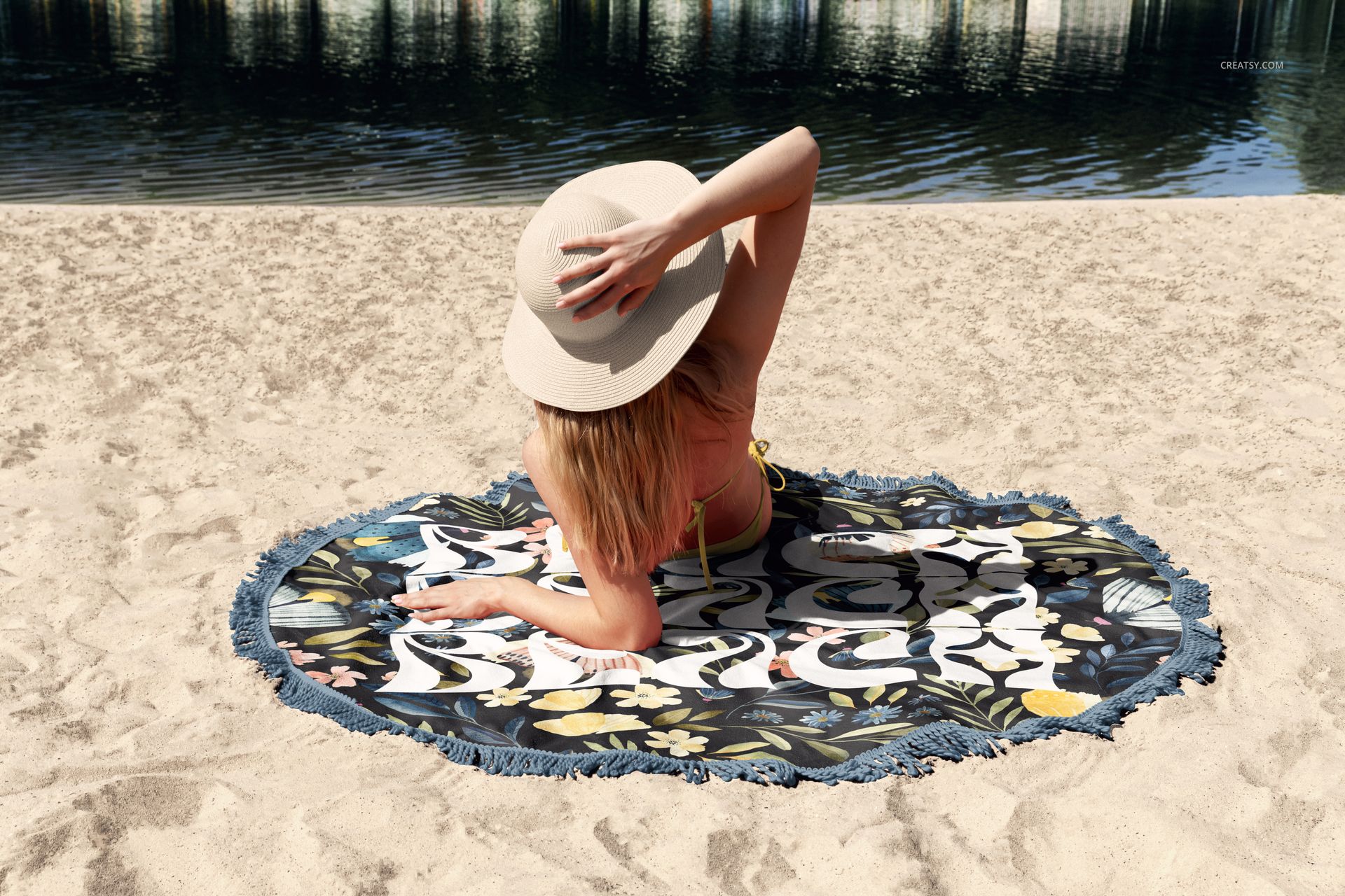 Top view of round beach towel mockup featuring blue fringe and colorful botanical design, placed on sand by water.