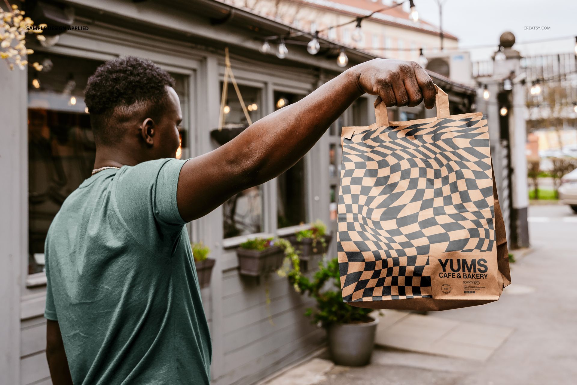 Outdoor scene with a hand holding a patterned paper bag, displaying a mockup for packaging design.
