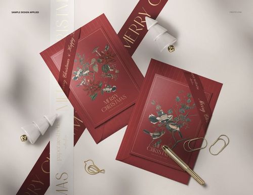 Christmas card PSD mockup featuring two red cards with floral designs, gold pen, envelopes, and holiday props on a table.