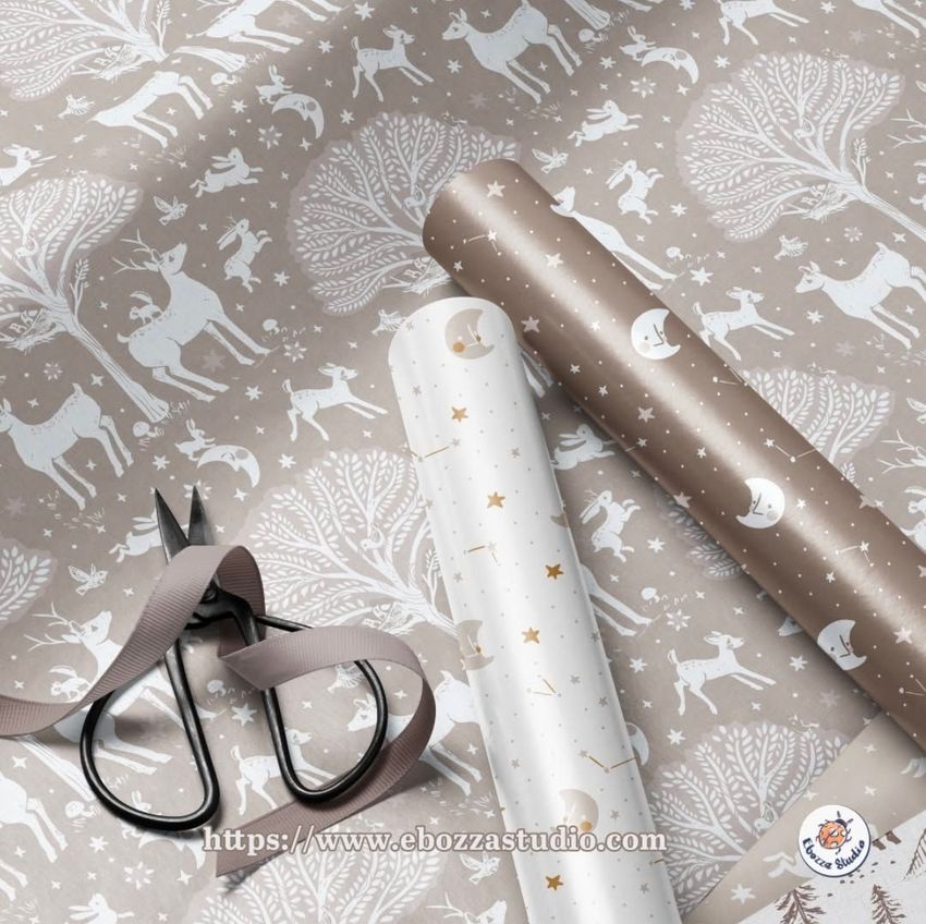 Gift wrapping paper mockup featuring two rolled sheets with woodland and celestial patterns, scissors, and ribbon on a flat surface.