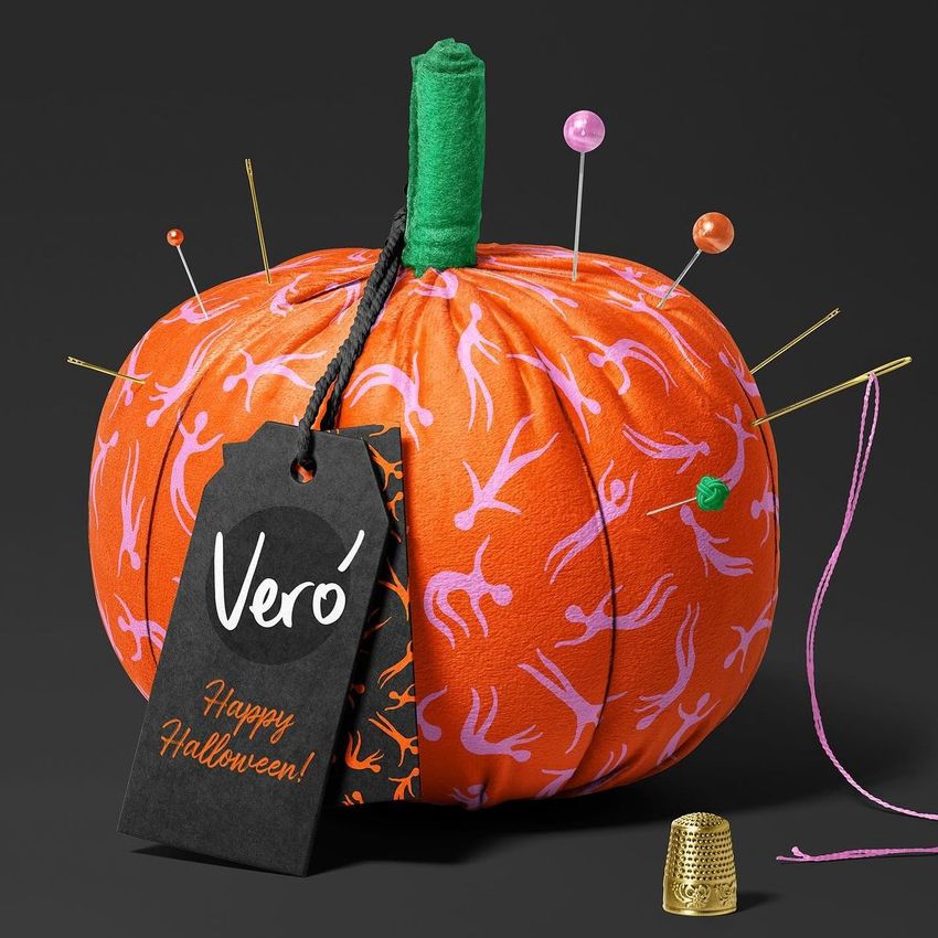 Fabric pumpkin mockup with orange fabric featuring pink patterns, used as a pincushion with pins and a black gift tag.