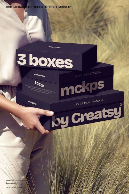 Person holding three stacked rectangular black boxes with bold white branding text, standing in tall grass outdoors.