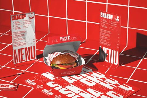 Front view of a fast food burger in a red branded box with striped wrapper, surrounded by red menus and coasters on a tiled surface.