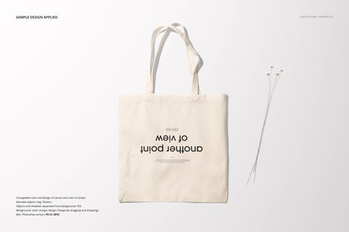 Front view of a plain beige tote bag mockup with black text, lying flat on a light background beside dried stems.