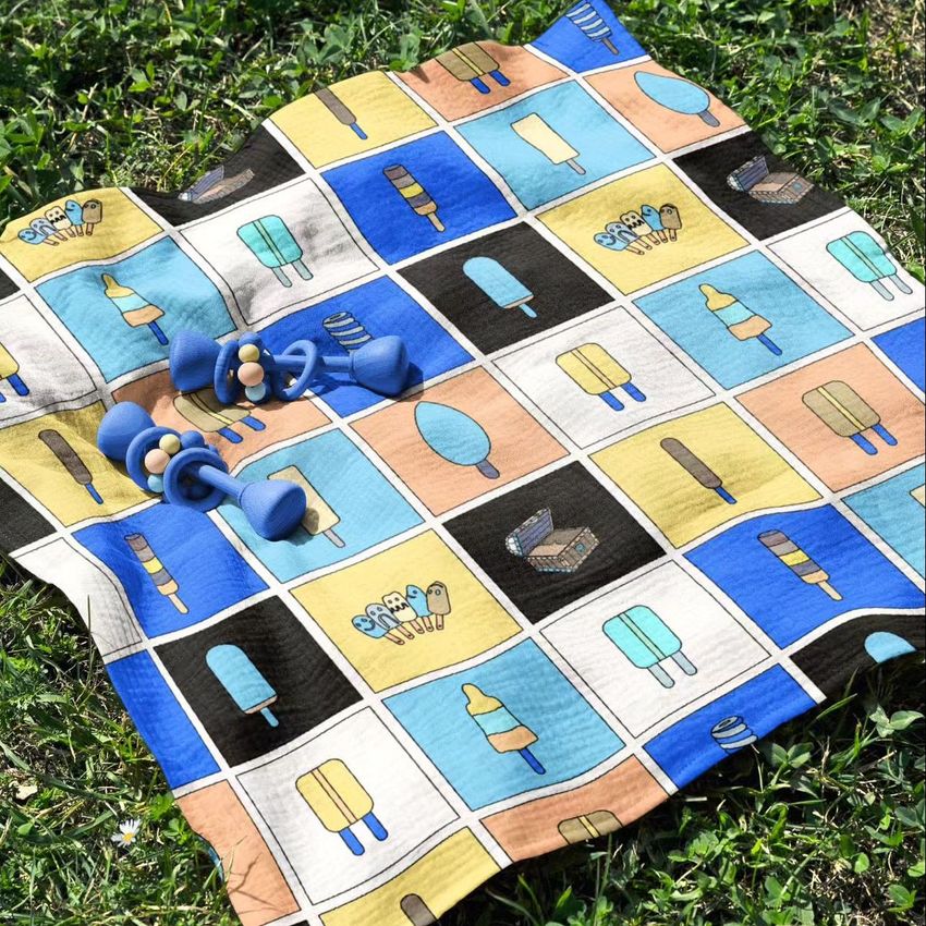 Crinkle cotton muslin swaddle blanket mockup featuring a colorful ice cream pattern, laid on green grass with blue baby rattles.