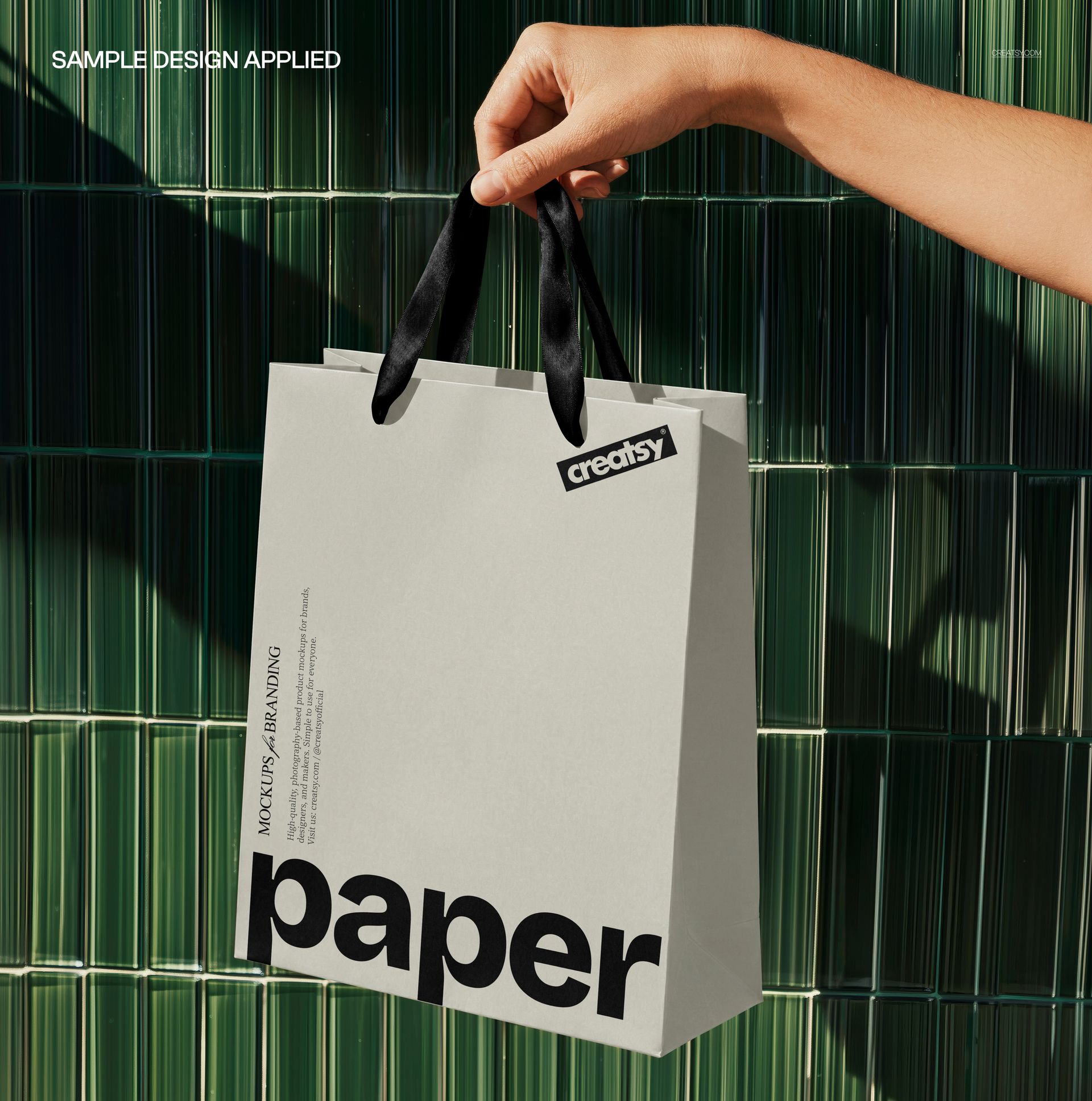 PSD mockup of a square-shaped shopping bag with black ribbon handles, held by a hand in sunlight near a green wall.