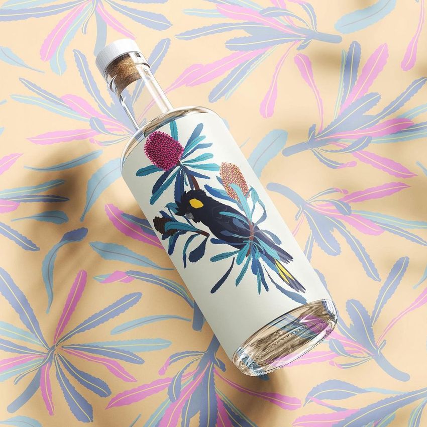 Glass bottle mockup with cork, featuring a black cockatoo and banksia illustration, placed on matching patterned paper background.