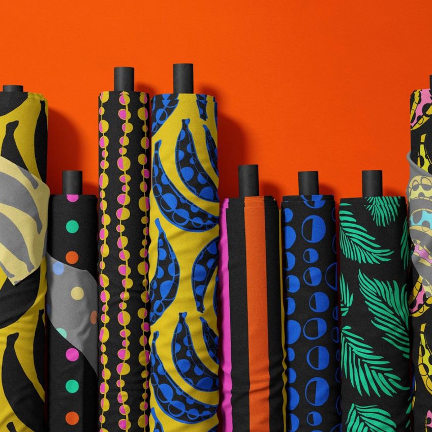 Fabric rolls mockup featuring nine upright rolls with bold, colorful patterns including bananas, dots, stripes, and leaves on a vivid orange background.