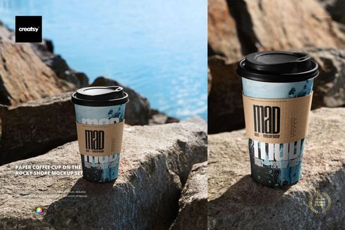 PSD mockup of a paper coffee cup with a black lid and custom sleeve placed among rocks beside a calm shoreline.