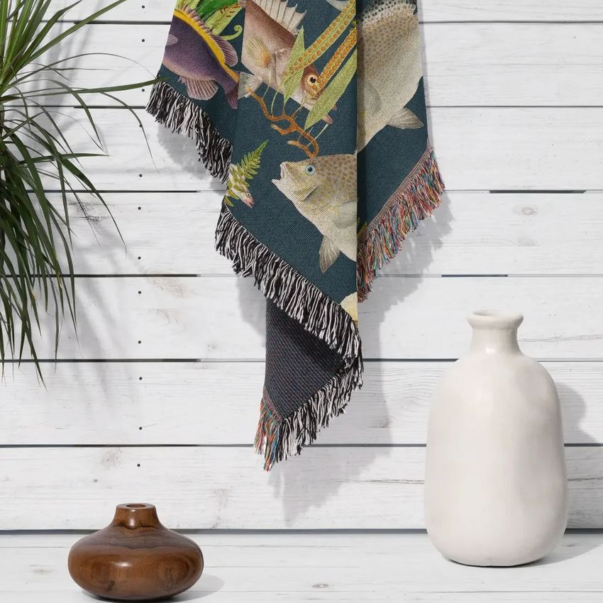 Woven throw blanket mockup with fringed edges, featuring colorful fish illustrations, draped against a white wood wall.