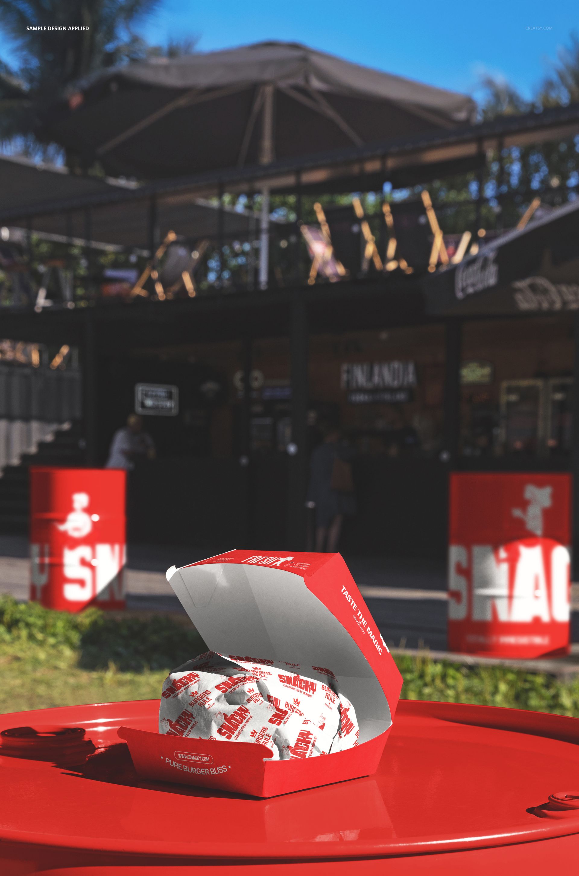 Red burger box mockup displayed on a barrel outdoors, with a wrapped burger inside and a food spot in the background.
