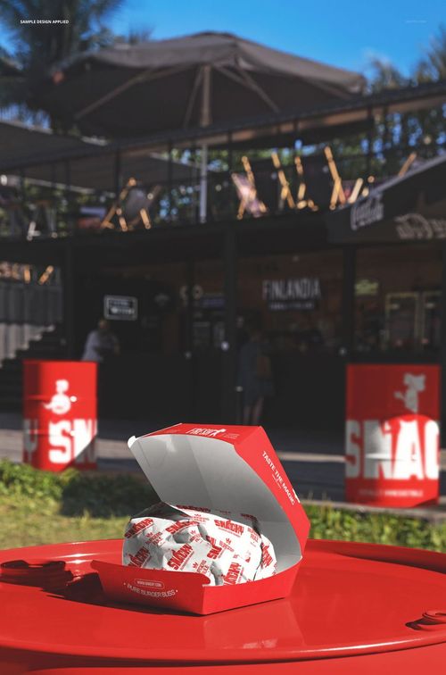Red burger box mockup displayed on a barrel outdoors, with a wrapped burger inside and a food spot in the background.