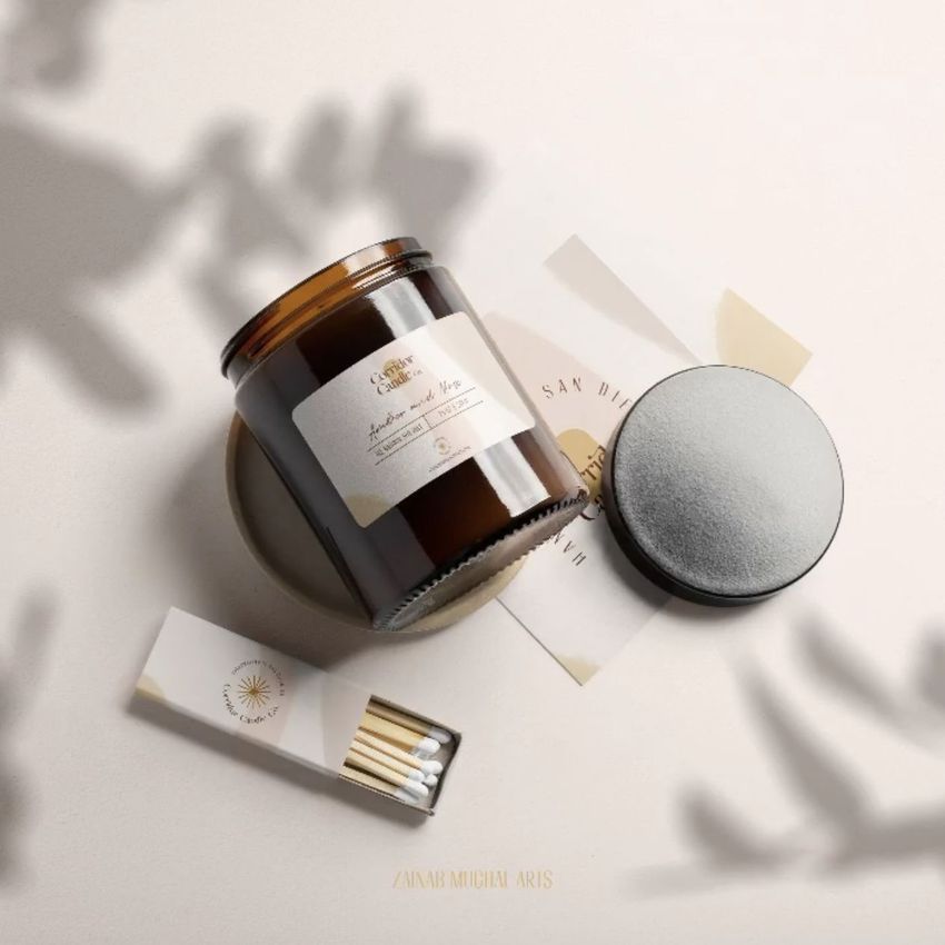 Sticker mockup featuring a brown glass candle jar with a label, matchbox, and paper cards arranged on a light surface.