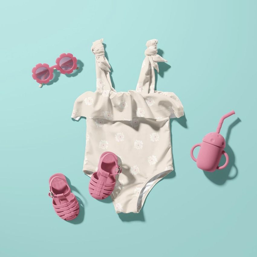 Baby swimsuit mockup featuring a light floral one-piece suit, pink sandals, flower-shaped sunglasses, and a sippy cup on a blue background.