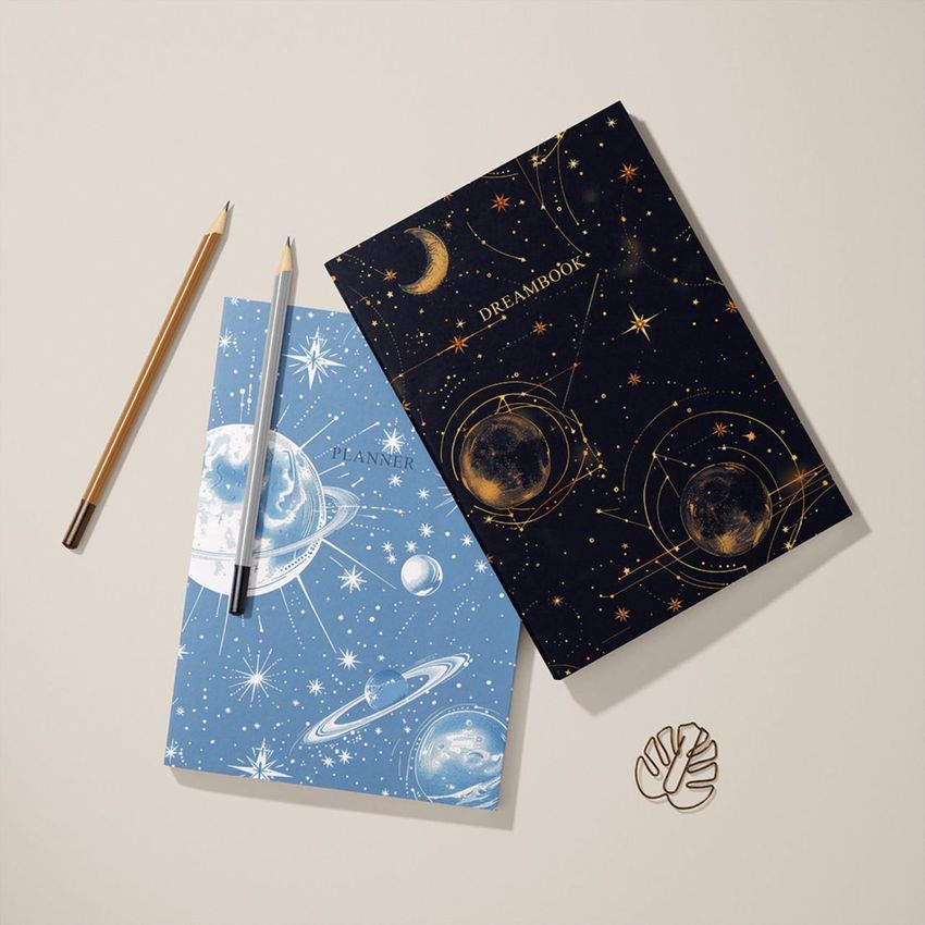 Top view of two planner mockup notebooks with celestial designs, one in blue and one in black, next to two pencils and a gold paper clip on a light surface.
