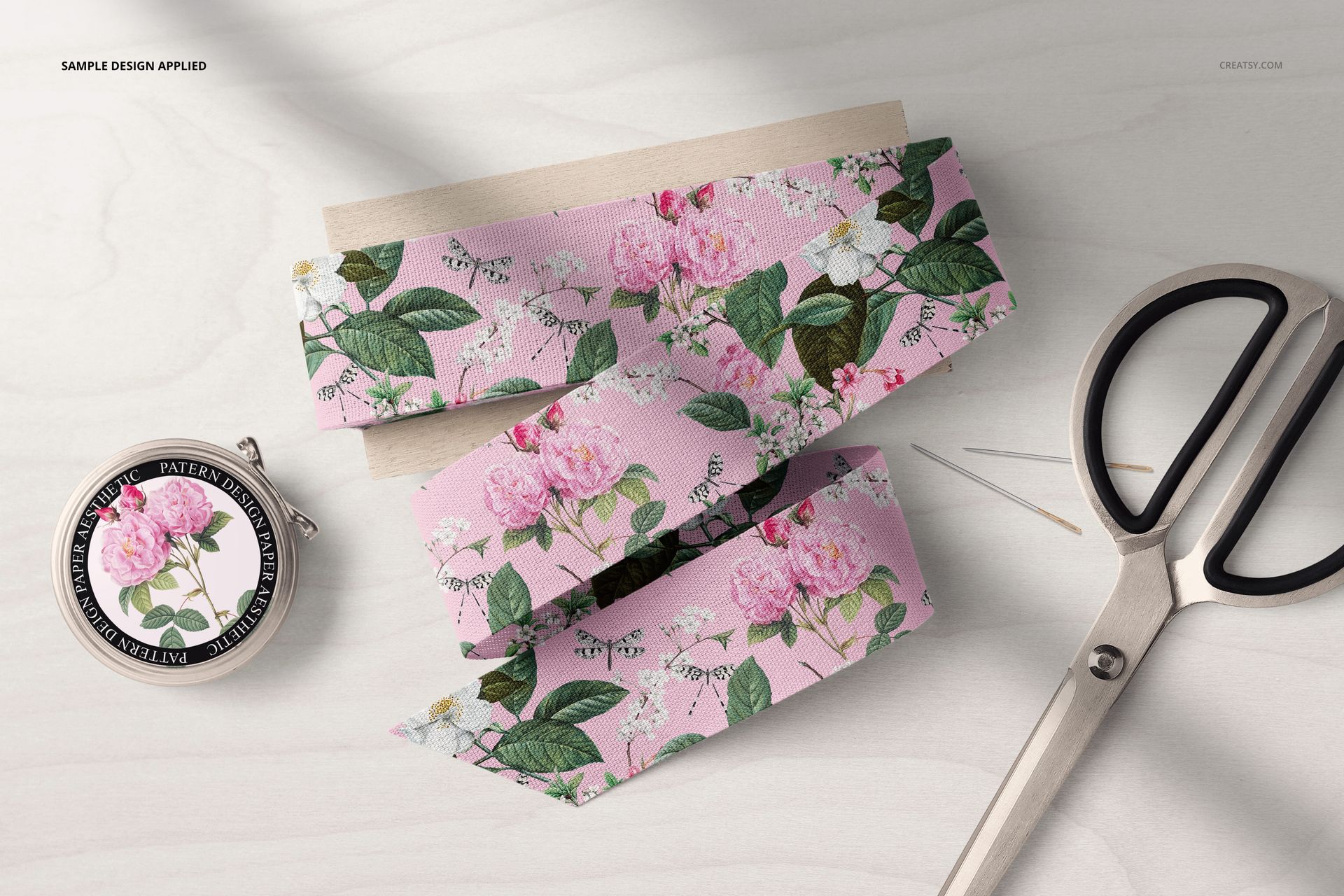 Cotton ribbon mockup with pink flowers and green leaves, sewing kit accessories, and scissors on a wooden background.