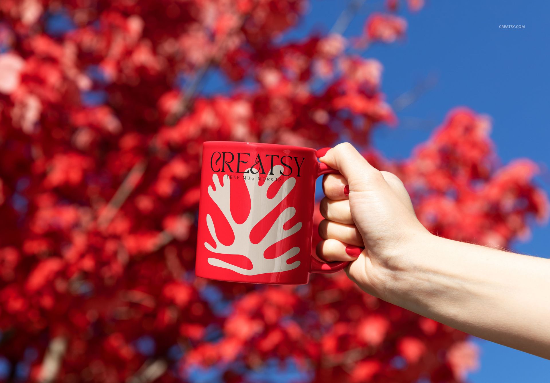 Outdoor PSD mockup showing a red coffee mug with white abstract pattern, held up near colorful foliage and sky.