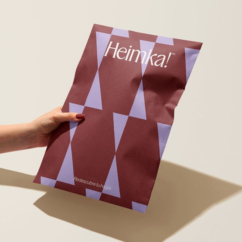 A hand holds a kraft mailer mockup with a geometric purple pattern and bold white text on a burgundy background, shown against a neutral backdrop.