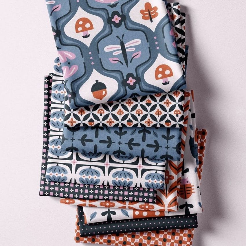 Fabric mockup featuring a stacked arrangement of folded textiles with whimsical forest-inspired patterns in blue, orange, and white tones.