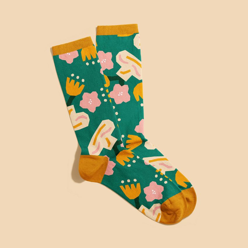 Socks mockup featuring a pair of green socks with bold yellow, pink, and white abstract floral patterns on a plain beige background.