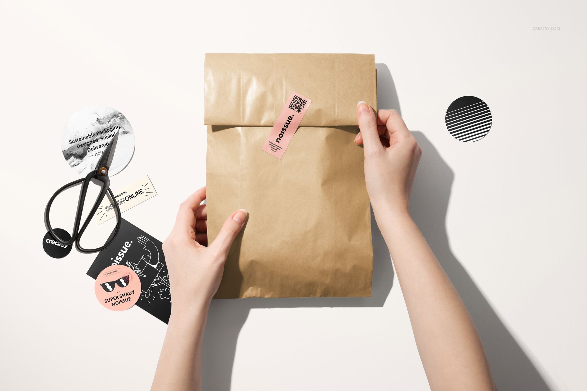 Top view of a paper bag mockup set, featuring hands, a pink sticker, and office supplies arranged on a white tabletop.