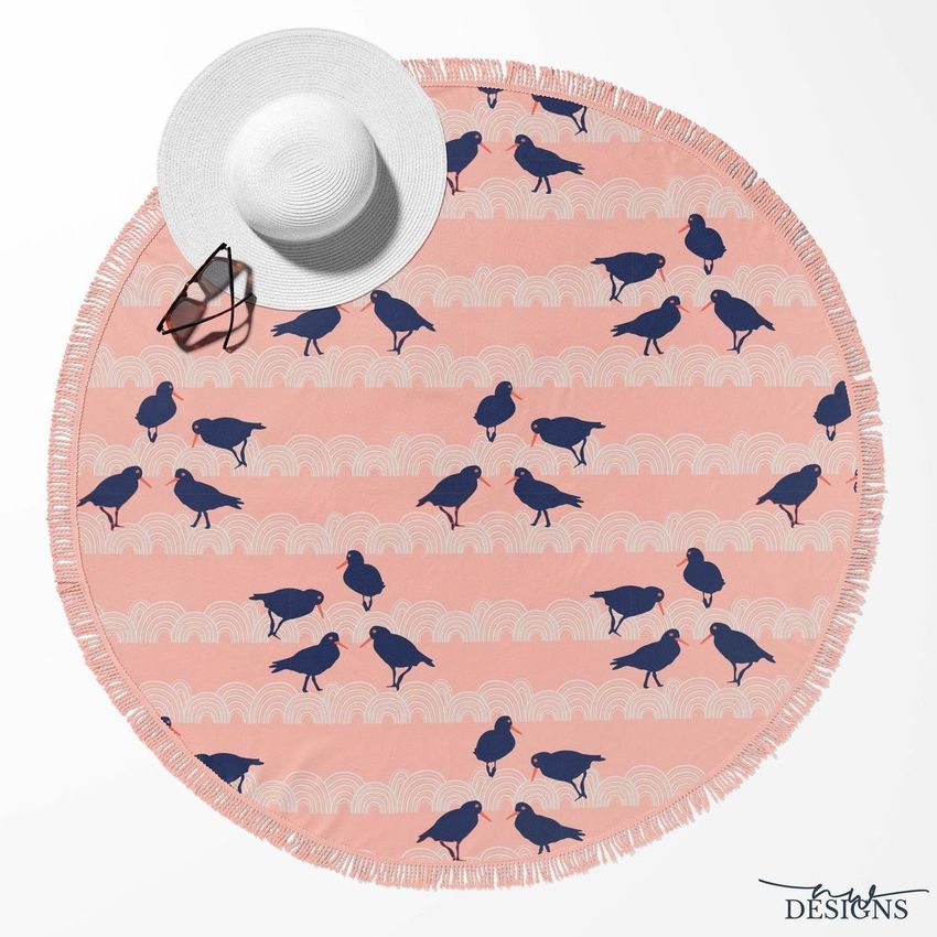 Round beach towel mockup with pink background, navy bird pattern, and tassel fringe, shown with a white sunhat and sunglasses.