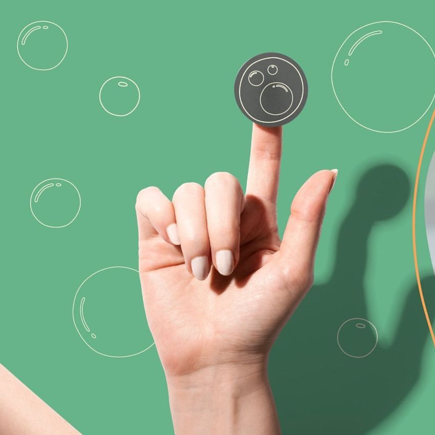 Hand with manicured nails holding a round sticker mockup featuring a simple face design on a green background with bubble graphics.