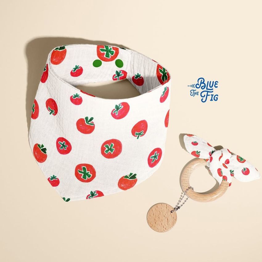 Baby muslin bandana mockup with a red tomato pattern, displayed next to a matching fabric and wood teether on a beige background.