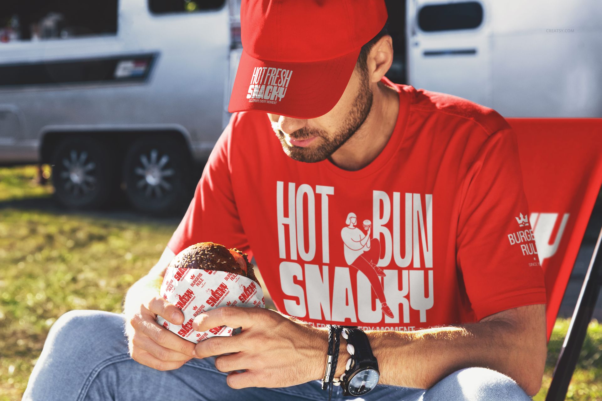 Person in red branded t-shirt and cap holding a wrapped burger outdoors, sitting on a red chair near a trailer.