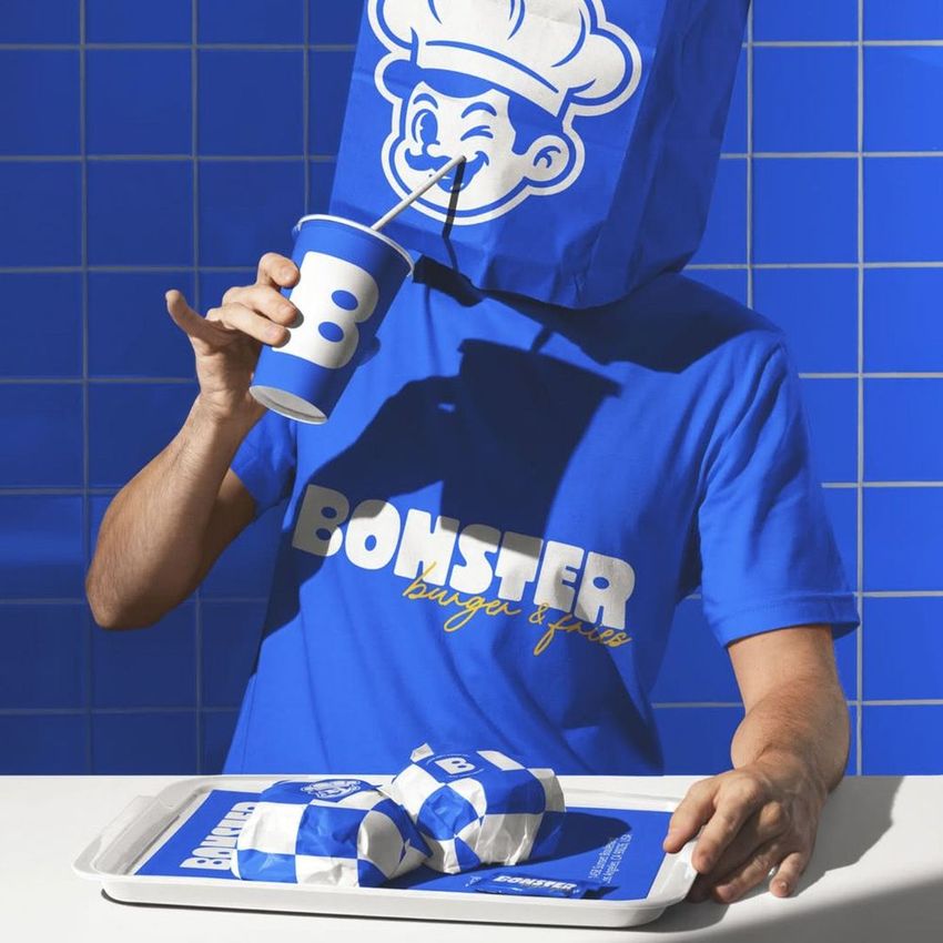 Person wearing a blue paper bag with a cartoon face sits at a table holding a branded fast food cup and tray with wrapped burgers.