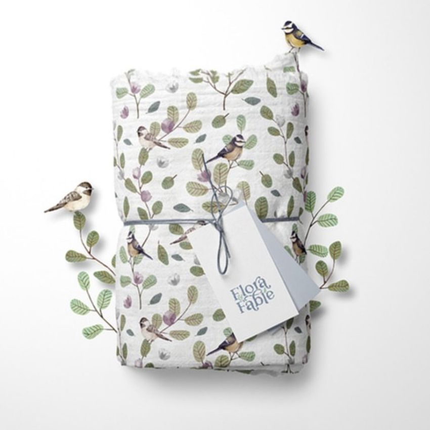 Muslin swaddle blanket mockup featuring a folded blanket with a bird and leaf pattern, tied with string and a tag, styled with illustrated birds and branches on a white background.