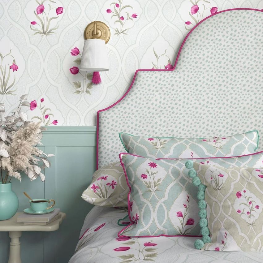 Bedroom pillows, a patterned headboard, and floral wallpaper in soft green and pink tones with a bedside table and wall lamp.