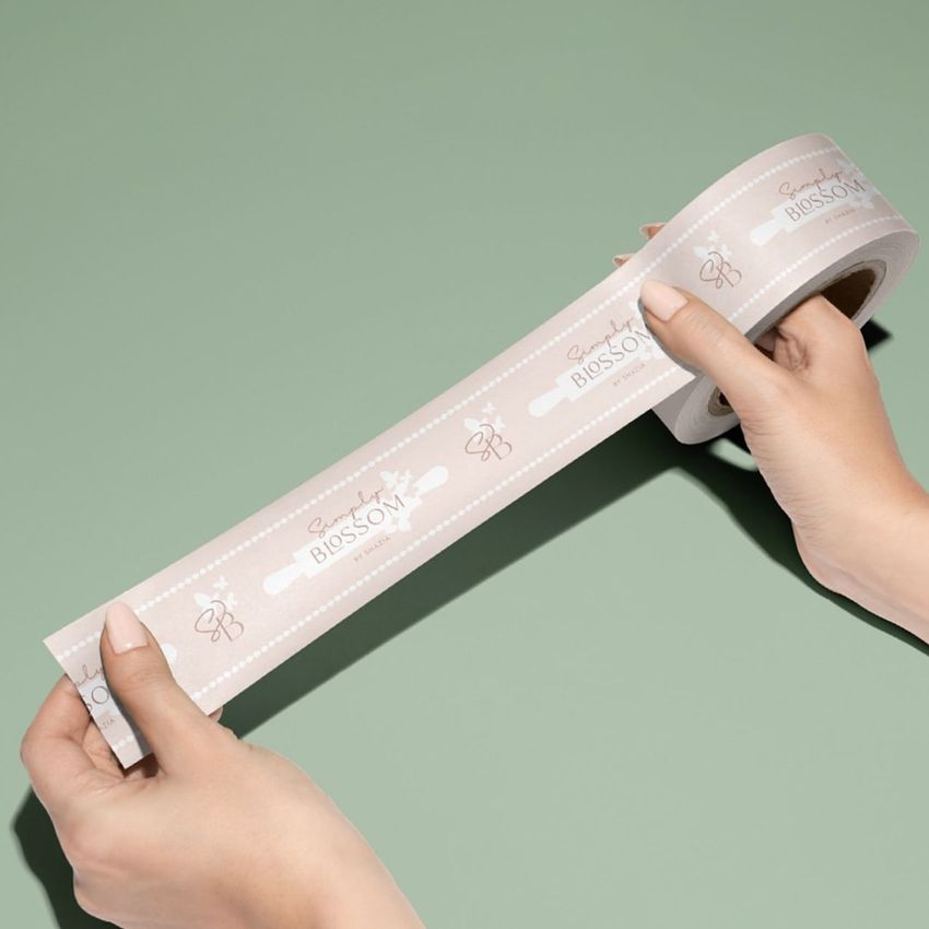 Two hands holding a roll of packing tape with a light beige background and repeated text and graphic elements, set against a muted green surface.