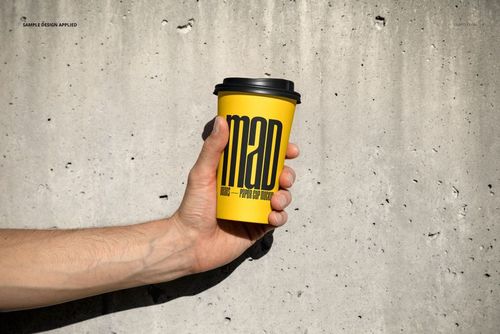 Front view of a yellow paper cup mockup with black lid and large black lettering, held by a hand near a gray wall.