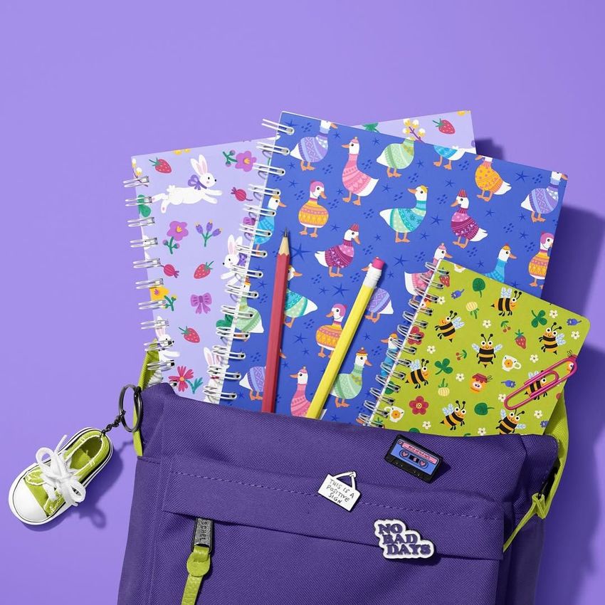 School backpack mockup with spiral notebooks featuring colorful animal and floral patterns, pencils, and a purple background.