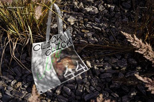 Mockup of a white tote bag with printed design, displayed on textured rocks and dry plants in natural sunlight.