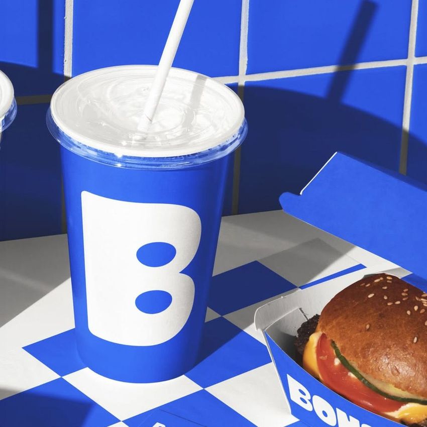 Burger branding mockup featuring a blue drink cup with a bold white letter, a burger in a branded box, and a blue checkered surface.