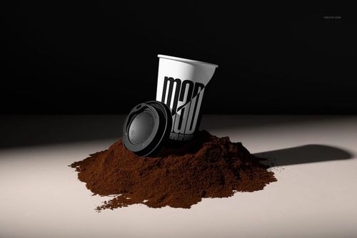PSD mockup of a white paper cup and black lid placed on a mound of ground coffee with dramatic lighting.