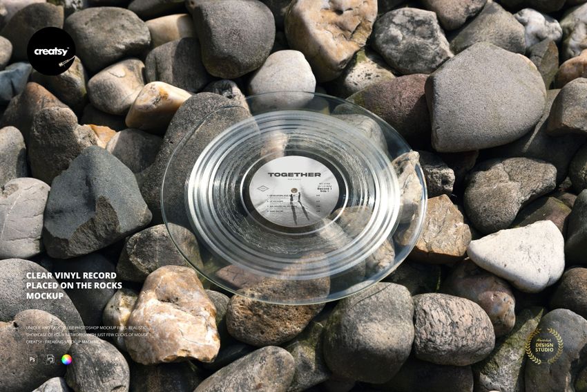 Clear vinyl record with visible grooves placed on a pile of smooth, multicolored rocks, top view mockup.