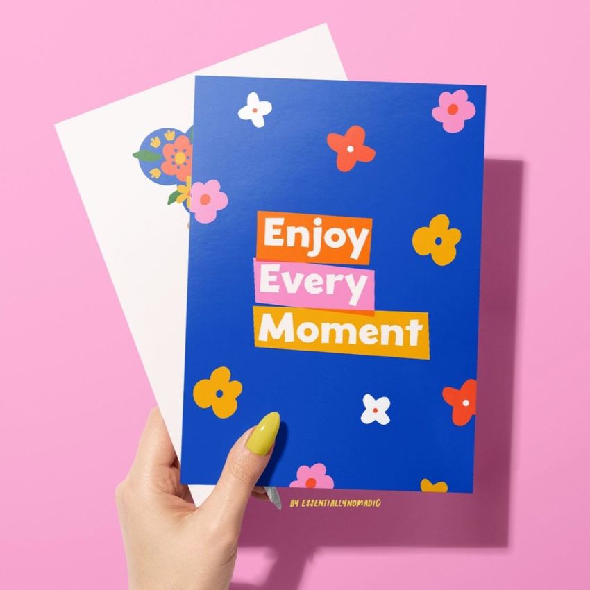 A woman's hand with yellow nail polish holds a greeting card mockup featuring bold text and colorful floral designs on a blue background.
