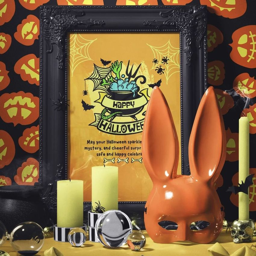 Halloween art print mockup featuring an ornate black picture frame with festive artwork, surrounded by yellow candles, pumpkins, and Halloween props.