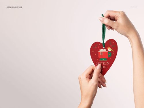 Close-up of two hands displaying a Christmas heart ornament with a green ribbon and holiday-themed design, PSD mockup.