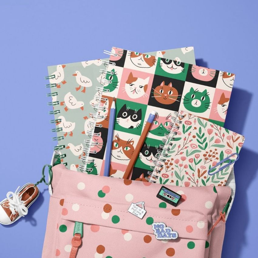 School backpack mockup with spiral notebooks featuring cat illustrations, floral patterns, and playful props on a purple background.