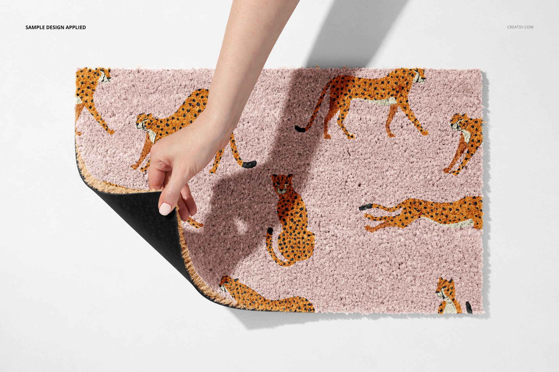 Rectangular door mat mockup featuring a pink surface with orange cheetah illustrations, shown from above with a hand folding the edge.