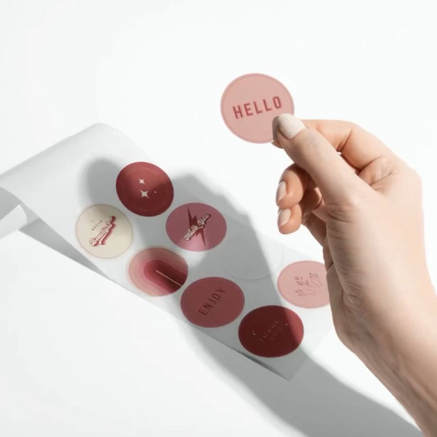Hand holding a round pink sticker with 'HELLO' text above a sheet of retro-themed stickers in various shades of red and beige.